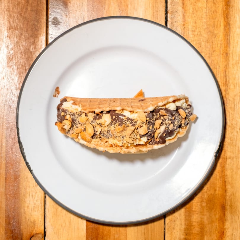 Waffle taco filled with chocolate, ice cream, and peanuts on a white plate.
