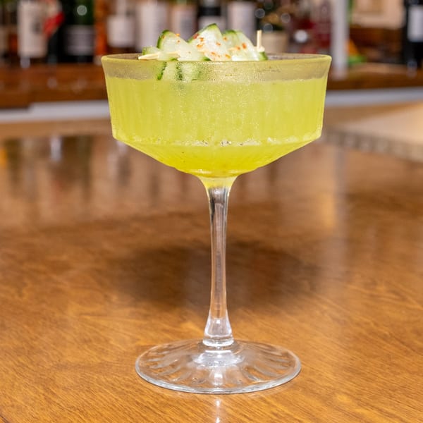 Bright green cocktail in a coupe glass, garnished with cucumber slices and spices on a wooden bar.
