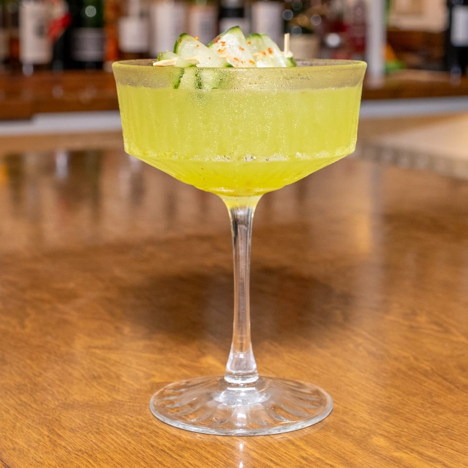 Bright green cocktail in a coupe glass, garnished with cucumber slices and spices on a wooden bar.