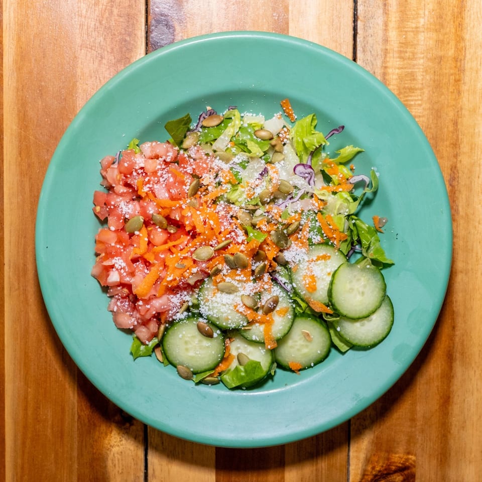 A vibrant salad with diced tomatoes, shredded carrots, sliced cucumbers, lettuce, and pumpkin seeds.