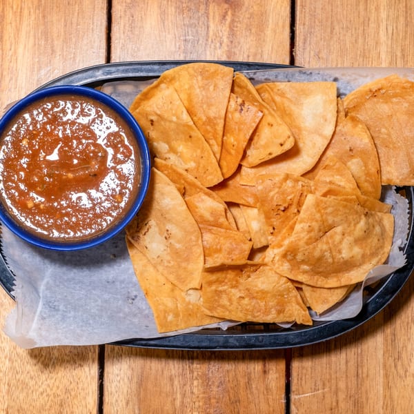 A blue bowl of chunky red salsa next to a basket of golden tortilla chips on a wooden table.