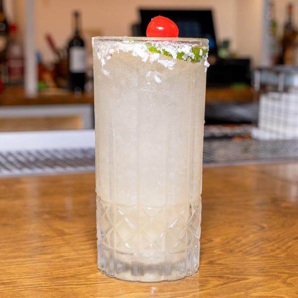 A tall, frosted cocktail with a salt rim and cherry garnish sits on a wooden bar.