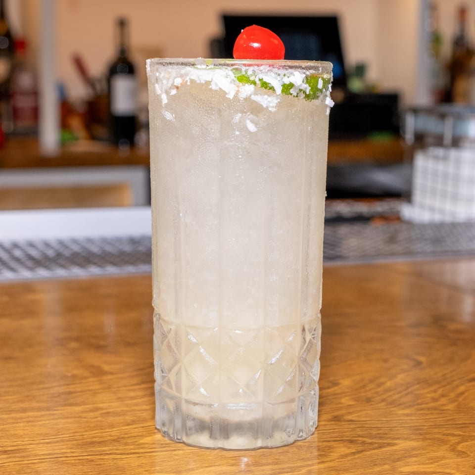 A tall, frosted cocktail with a salt rim and cherry garnish sits on a wooden bar.