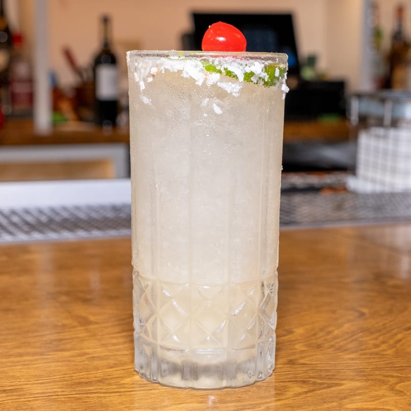 A tall, frosted cocktail with a salt rim and cherry garnish sits on a wooden bar.