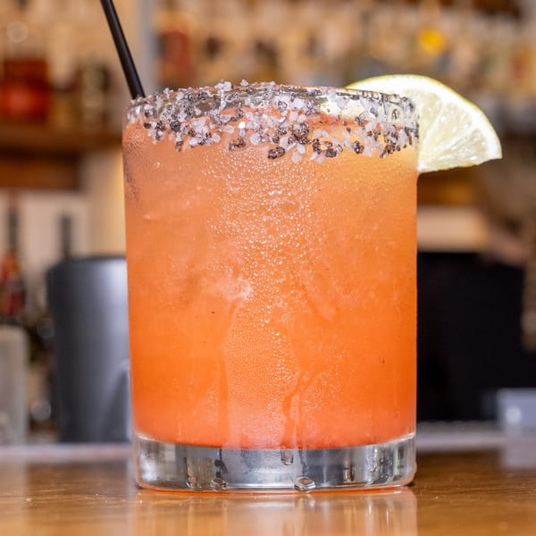 A vibrant orange cocktail with a salted rim and lime wedge, on a wooden bar.